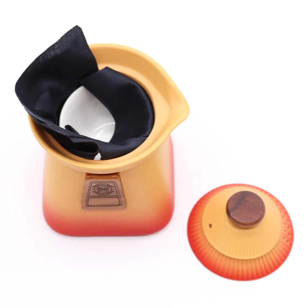 Tetera de cerámica naranja y roja, con tapa de madera y tela negra en el interior, sobre un fondo blanco.