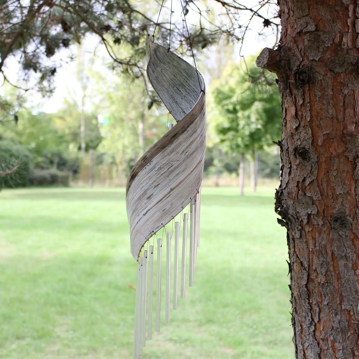 Campanilla de viento de hoja de coco blanqueada con forma espiral y tubos metálicos colgando en un jardín.