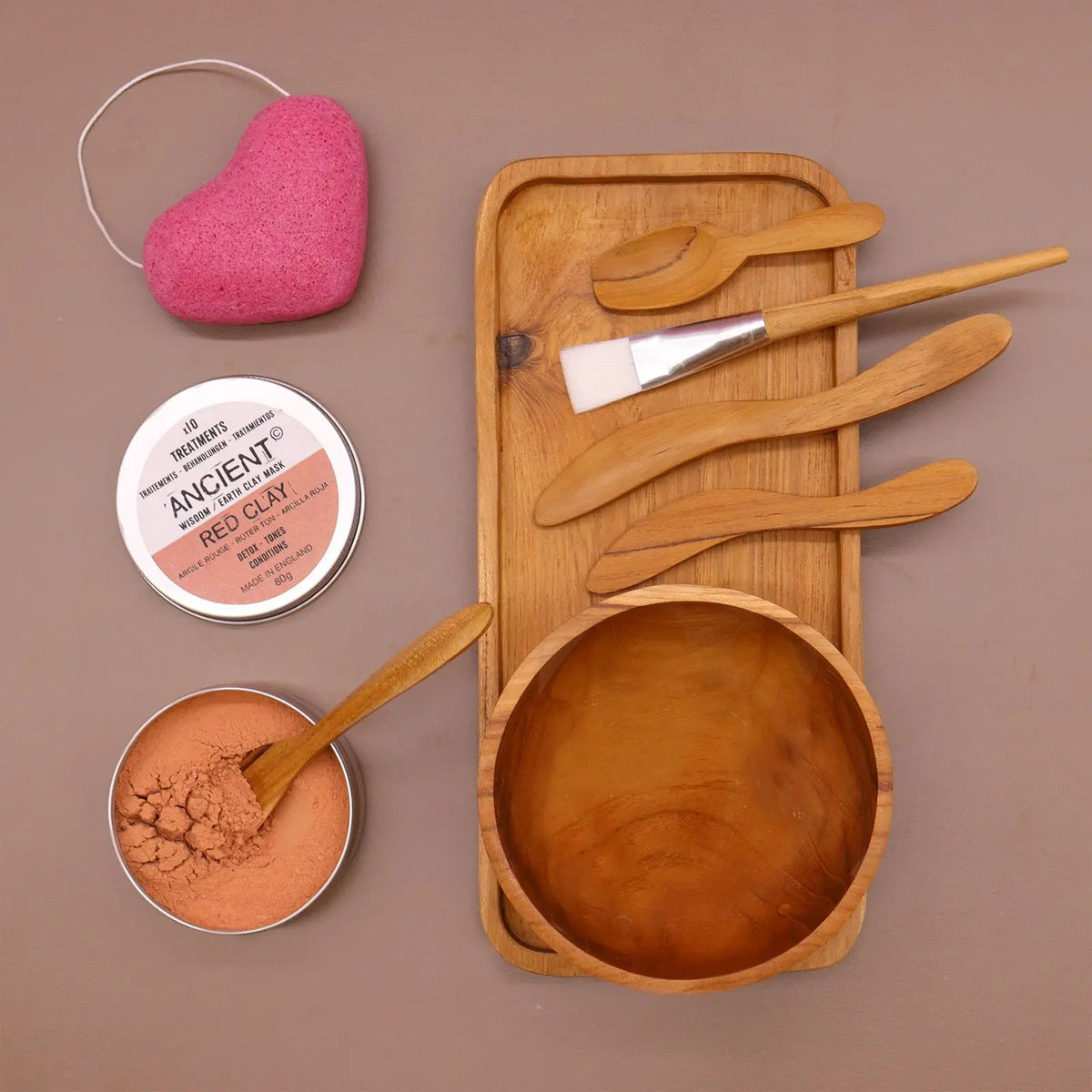 Kit de spa con cuenco, bandeja, espátulas de madera, brocha, esponja rosa y arcilla en envase metálico sobre fondo beige.