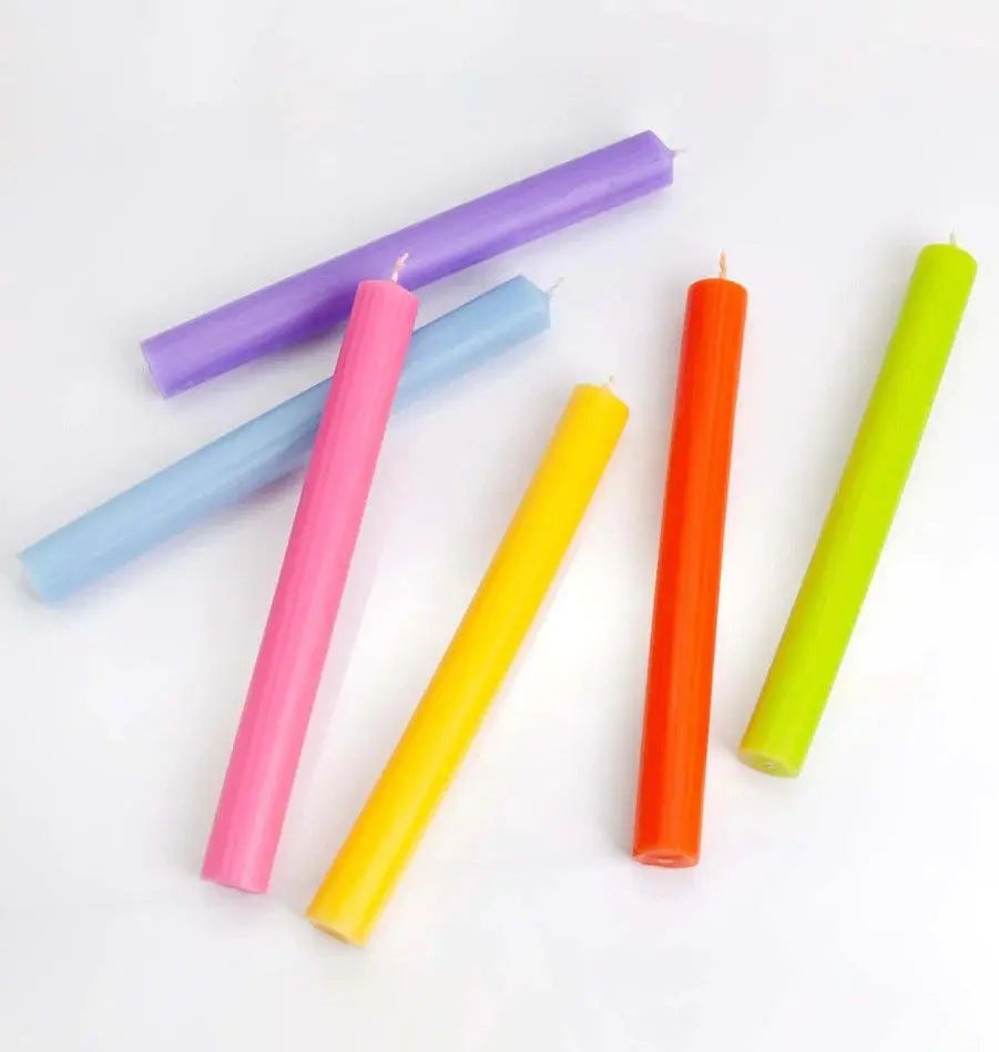 Velas cilíndricas largas de colores vivos (lila, azul, rosa, amarillo, naranja y verde) sobre un fondo blanco liso.