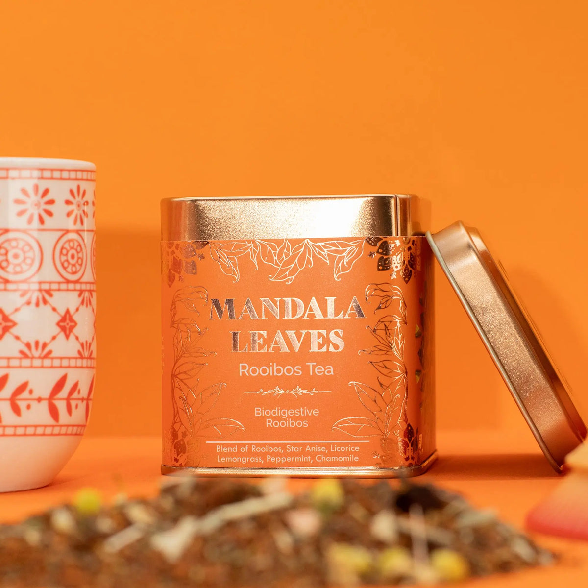 Lata metálica cuadrada naranja con detalles dorados, junto a una taza decorada y té a granel sobre fondo naranja.