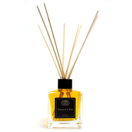 Difusor de vidrio cuadrado con líquido amarillo y varillas de madera, sobre fondo blanco. Etiqueta negra al frente.