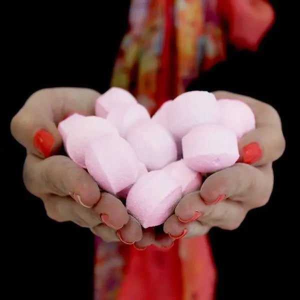 Manos sosteniendo un puñado de mini bombas de baño efervescentes de color rosa pálido con forma de pastilla.