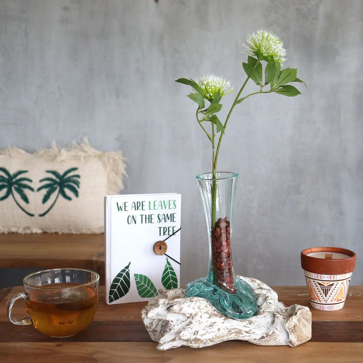 Florero de vidrio fundido sobre base de madera blanqueada, con flores blancas, sobre una mesa con objetos decorativos.