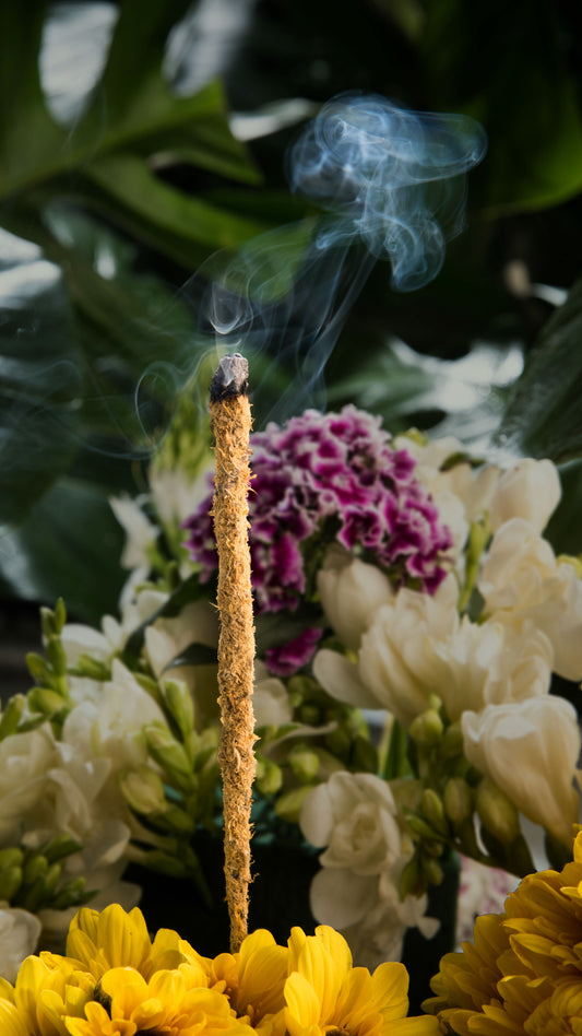 Incienso artesanal botánico de textura fibrosa emitiendo humo sobre un fondo de flores blancas, moradas y amarillas.