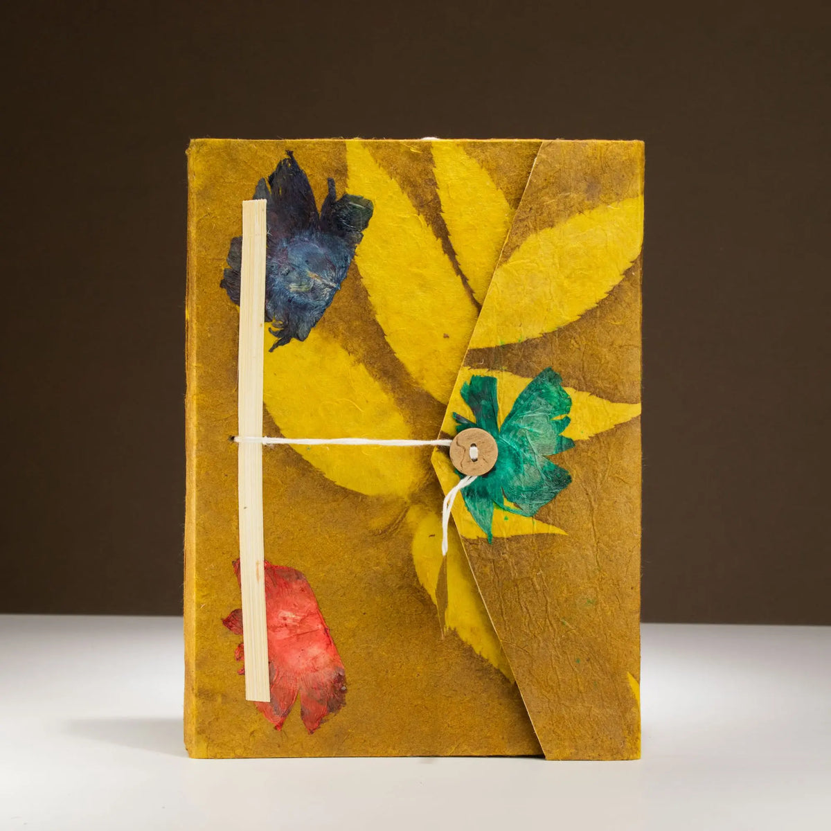 Cuaderno artesanal amarillo con flores azules y rojas, atado con cuerda, estilo bohemio y material ecológico.