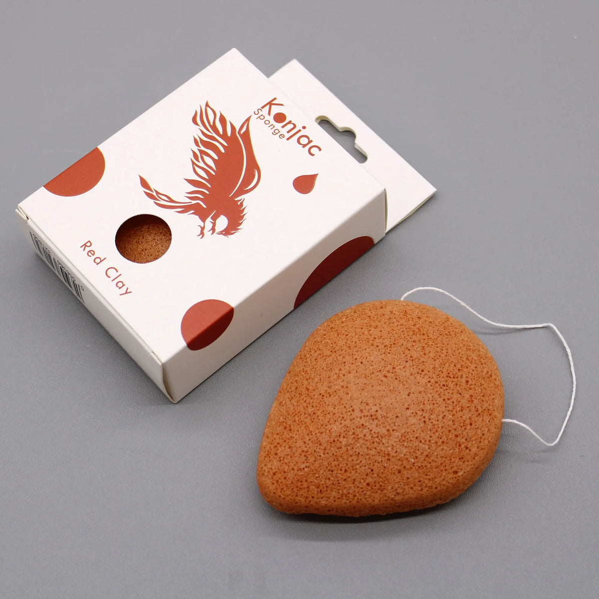 Esponja facial konjac de arcilla roja, forma de gota, textura porosa, con cordón blanco junto a su caja de empaque.