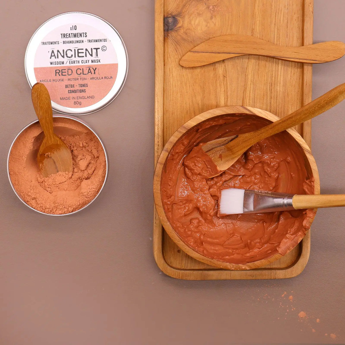 Set de spa con arcilla roja, cuenco y bandeja de madera de teca, espátulas y brocha de aplicación sobre fondo marrón.
