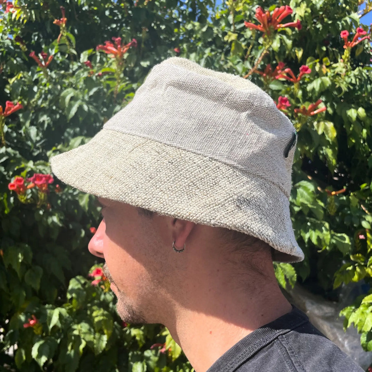 Sombrero tipo pescador de color natural, tejido rústico de cáñamo y algodón, usado por un hombre ante plantas con flores.