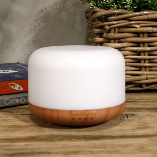 Difusor Aarhus blanco y madera, cilíndrico, con luz LED y temporizador, sobre mesa de madera junto a una cesta.
