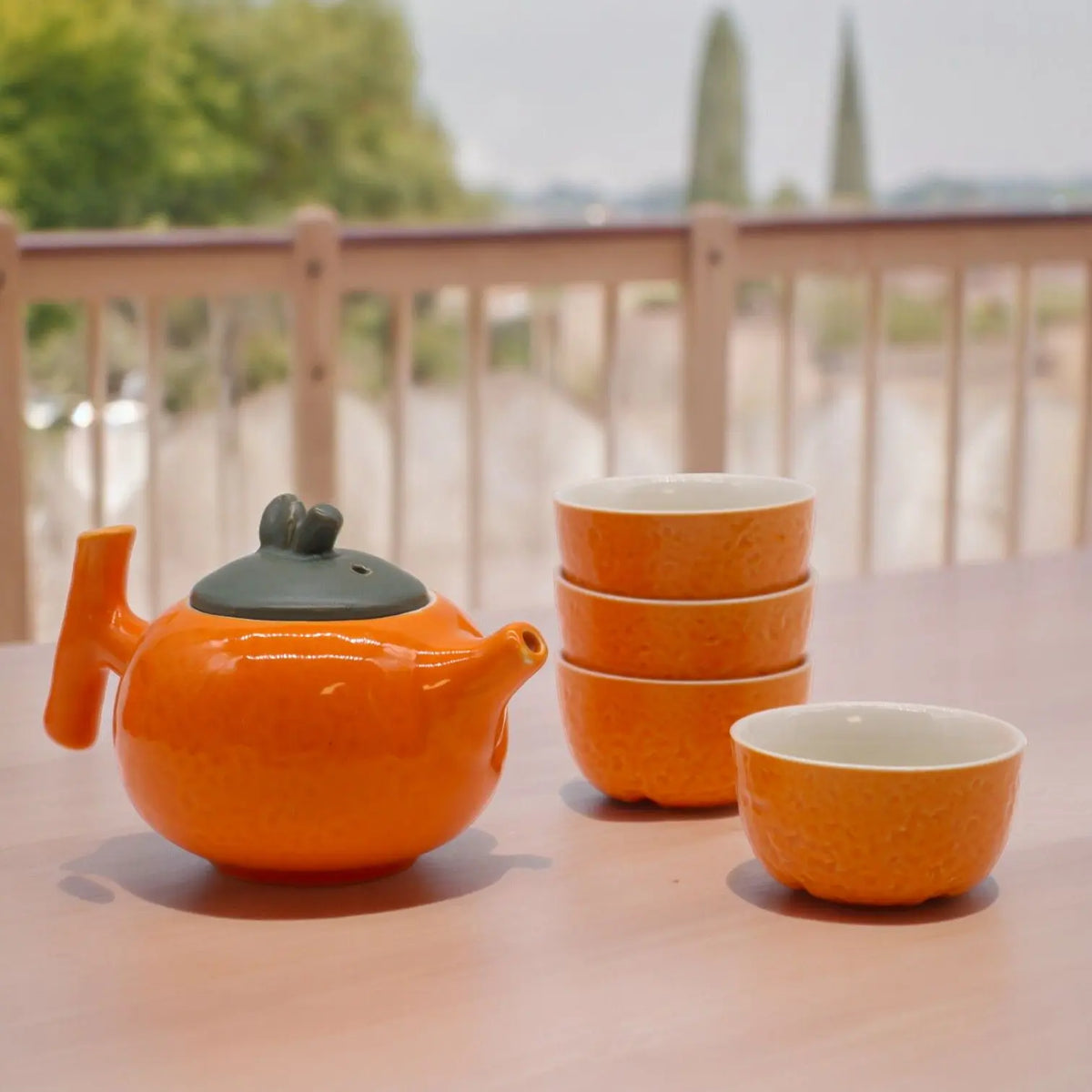 Tetera y tazas de cerámica naranja con textura rugosa, tapa gris, sobre una mesa al aire libre.