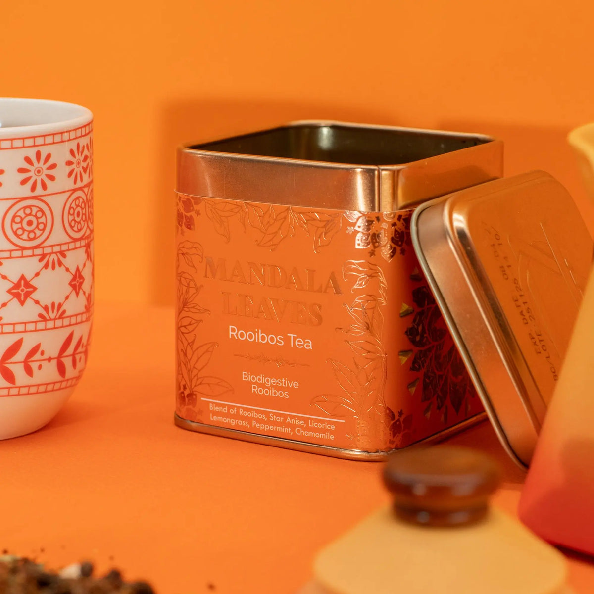 Lata metálica cuadrada de color naranja con té rooibos, junto a una taza decorada sobre fondo naranja.