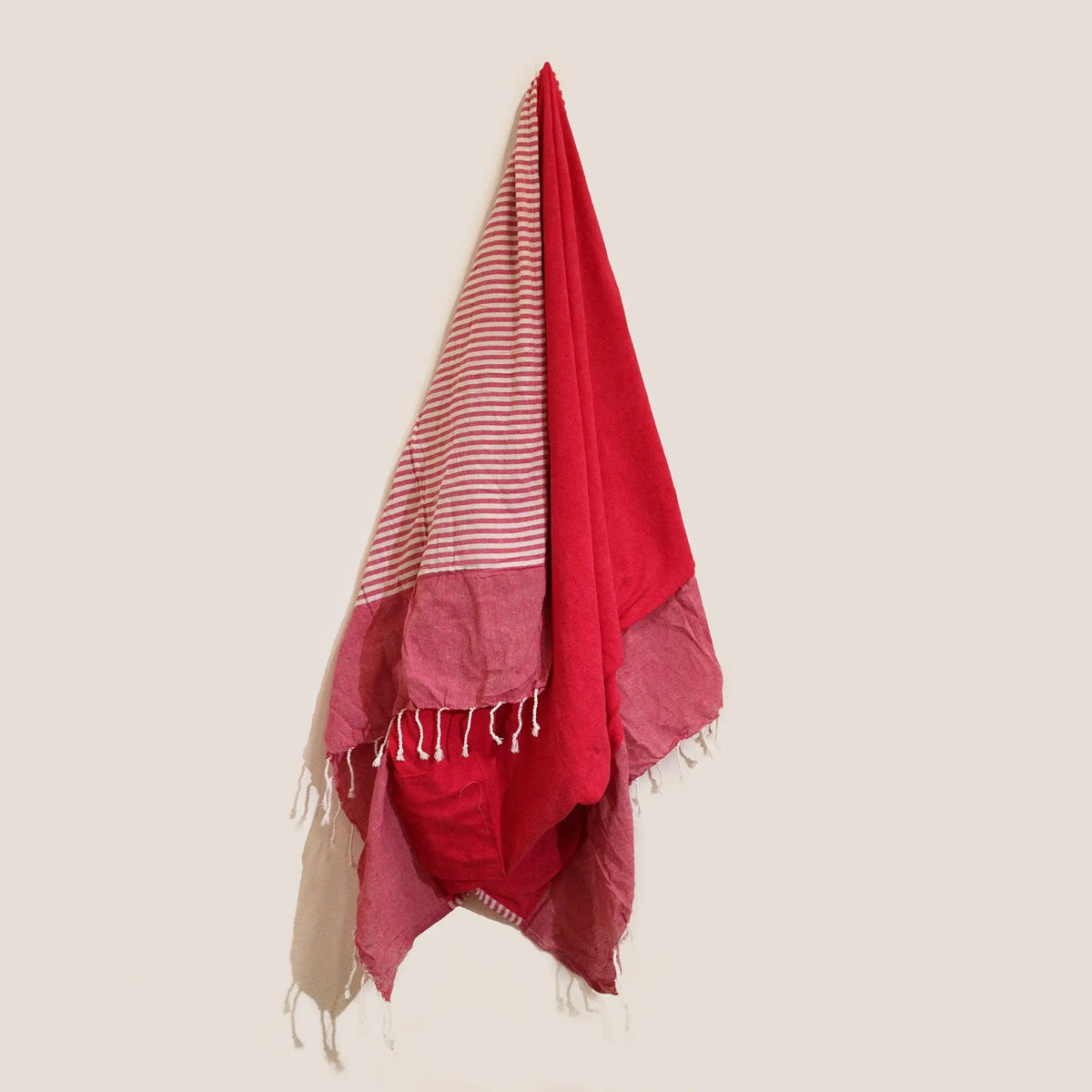 Toalla pareo de algodón rosa, con rayas y flecos, colgada verticalmente sobre un fondo de color crema.