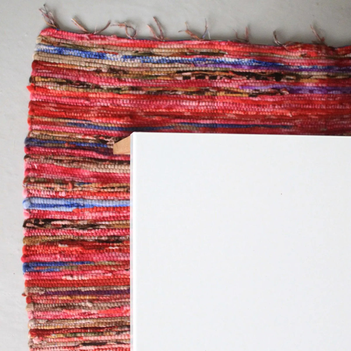 Alfombra de trapo tejida en tonos rojos, multicolores y flecos, parcialmente cubierta por una superficie blanca.