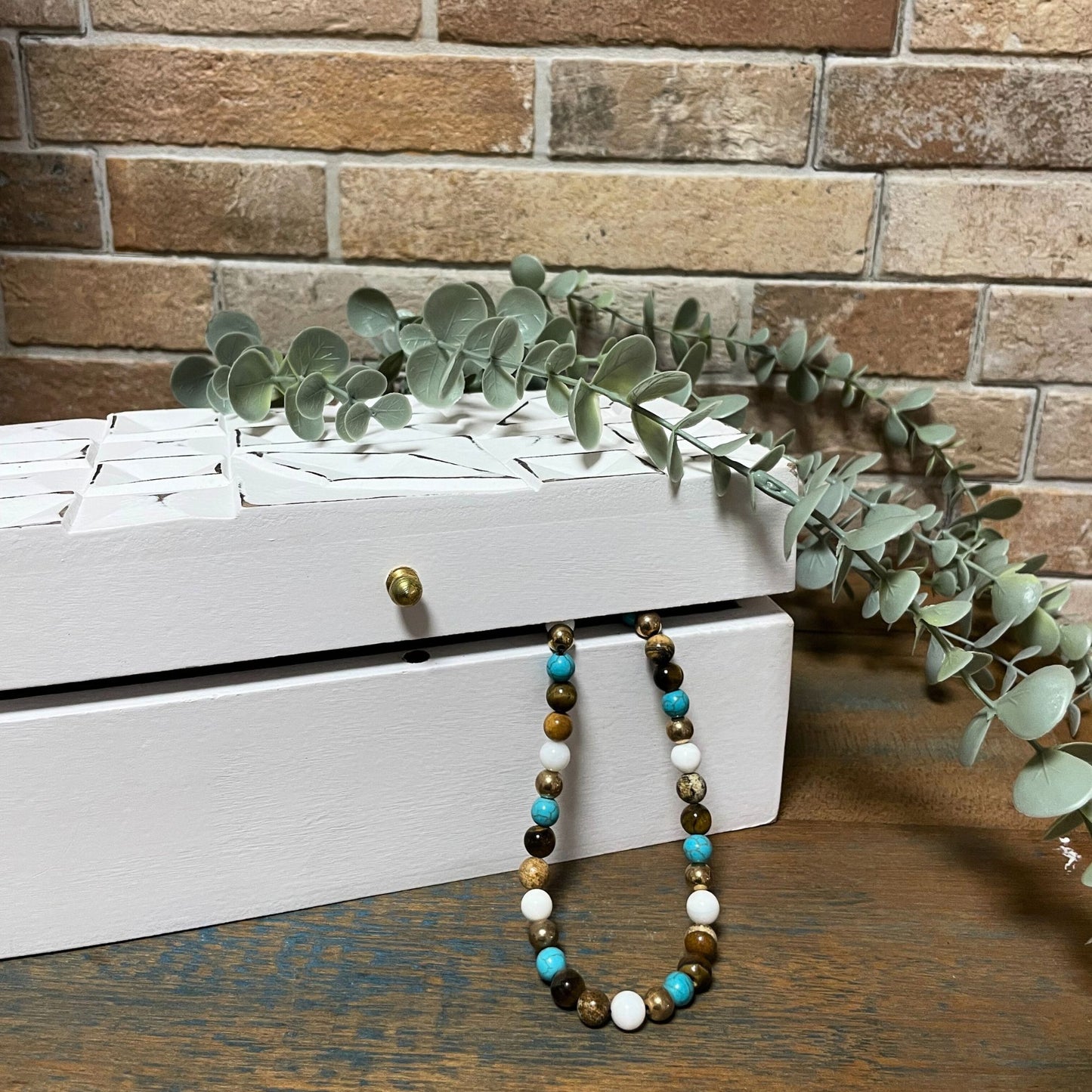 Collar de piedras naturales en tonos azules, marrones y blancas, sobre caja blanca decorativa, con fondo de ladrillo.