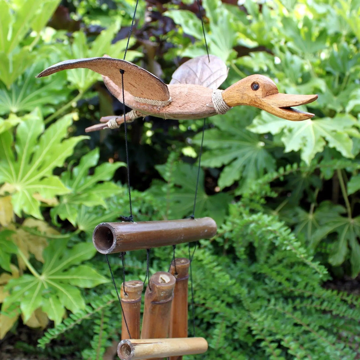 Carrillón de viento artesanal con figura de pato tallado en madera sobre tubos de bambú, colgado en un jardín verde.