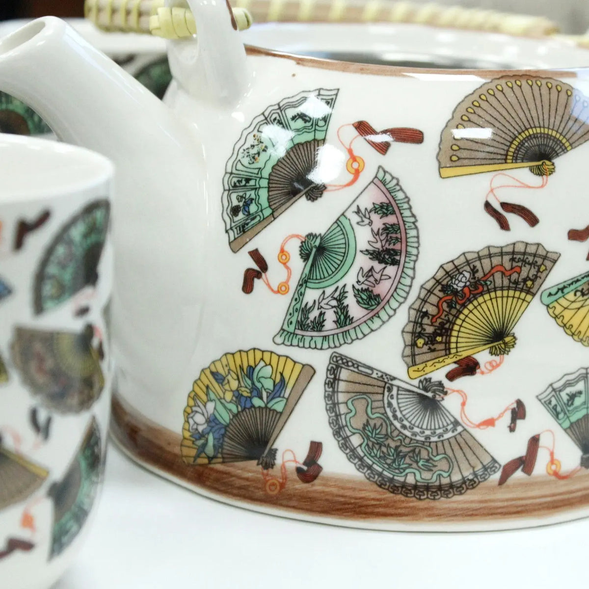 Tetera de cerámica blanca y taza con estampado de abanicos chinos coloridos y mango de mimbre.