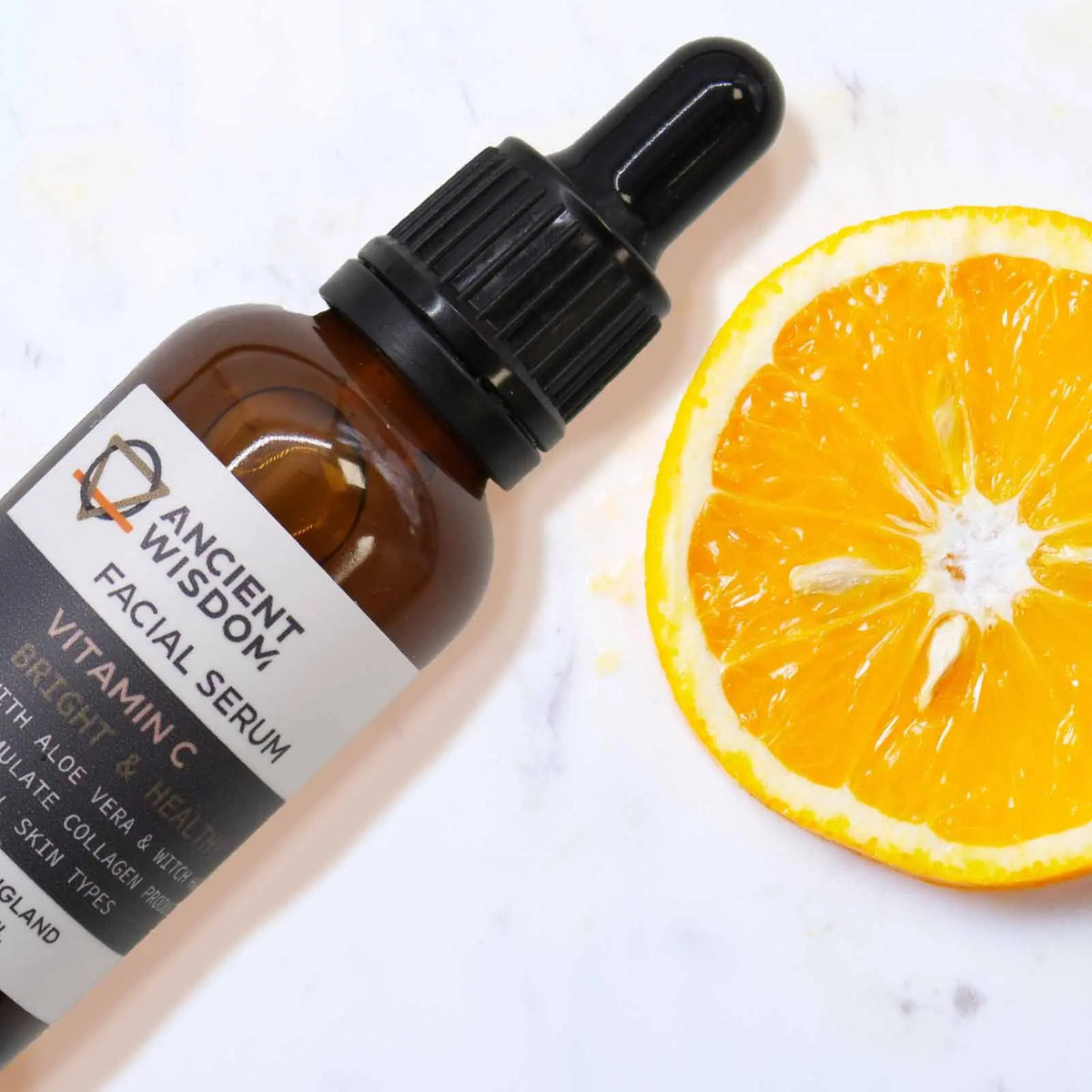 Serum facial de Vitamina C Ancient Wisdom en frasco ámbar con gotero, junto a una rodaja de naranja sobre fondo claro.