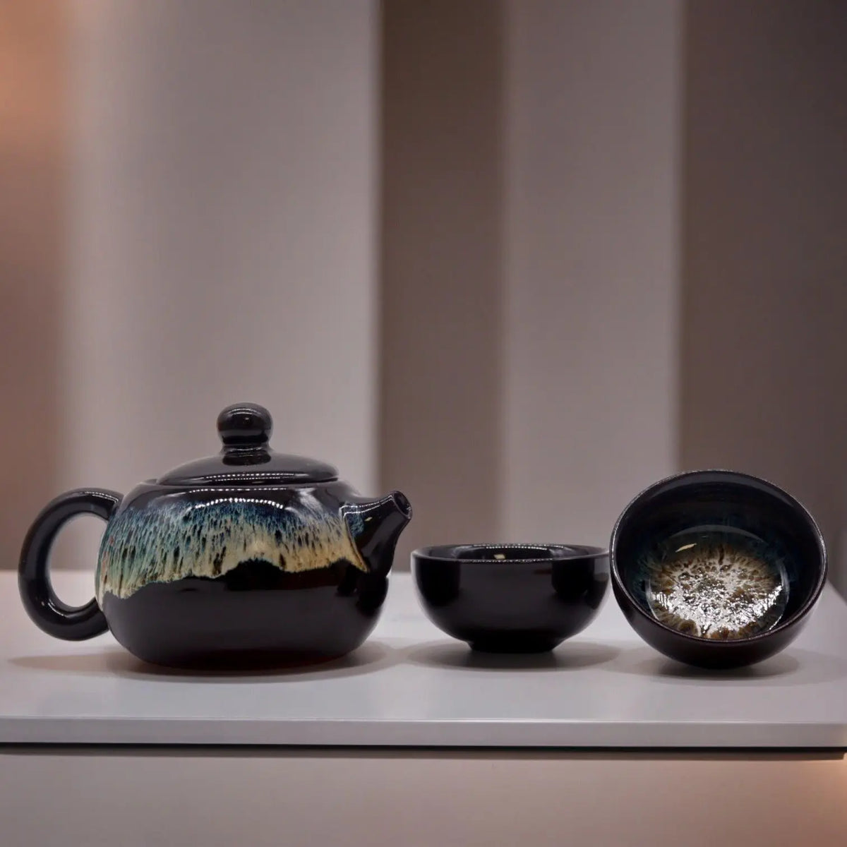 Set de té esmaltado negro y azul con tetera y dos tazas de cerámica de diseño artesanal sobre superficie blanca.