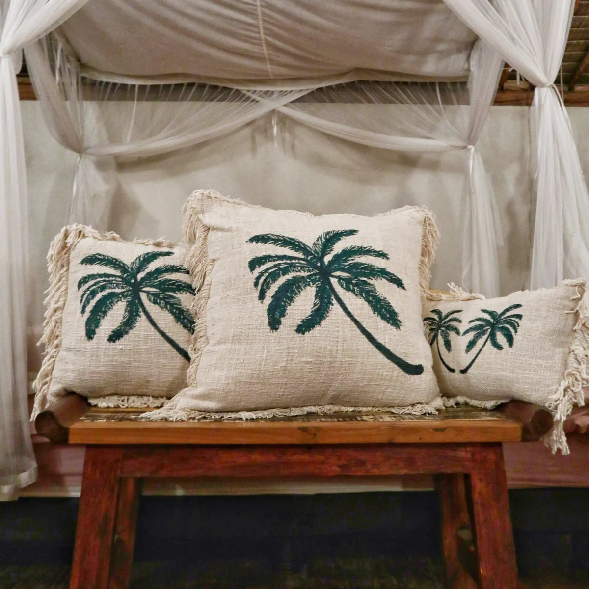 Funda de cojín de lino beige con estampado de palmera verde oscuro y flecos, sobre un banco de madera.