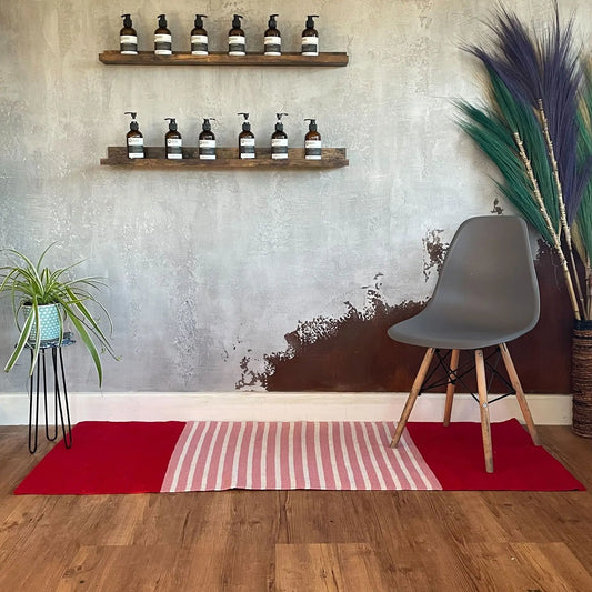 Alfombra rectangular de algodón rojo y rosa, con secciones sólidas y a rayas, sobre suelo de madera.