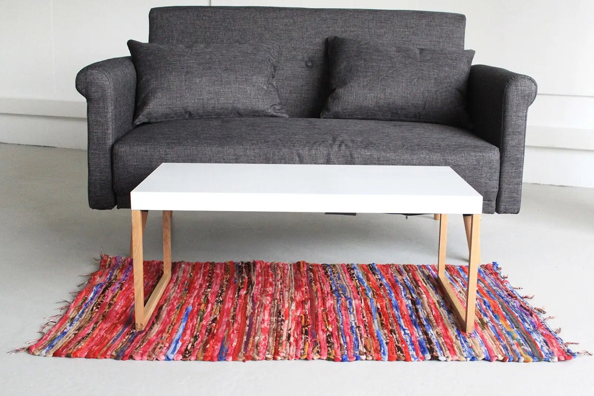 Alfombra de trapo artesanal con rayas multicolores texturizadas bajo una mesa blanca frente a un sofá gris