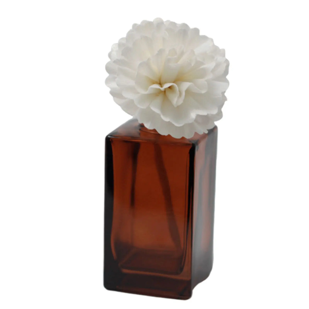 Flor difusora blanca de papel sobre frasco de vidrio ámbar rectangular. Fondo blanco.