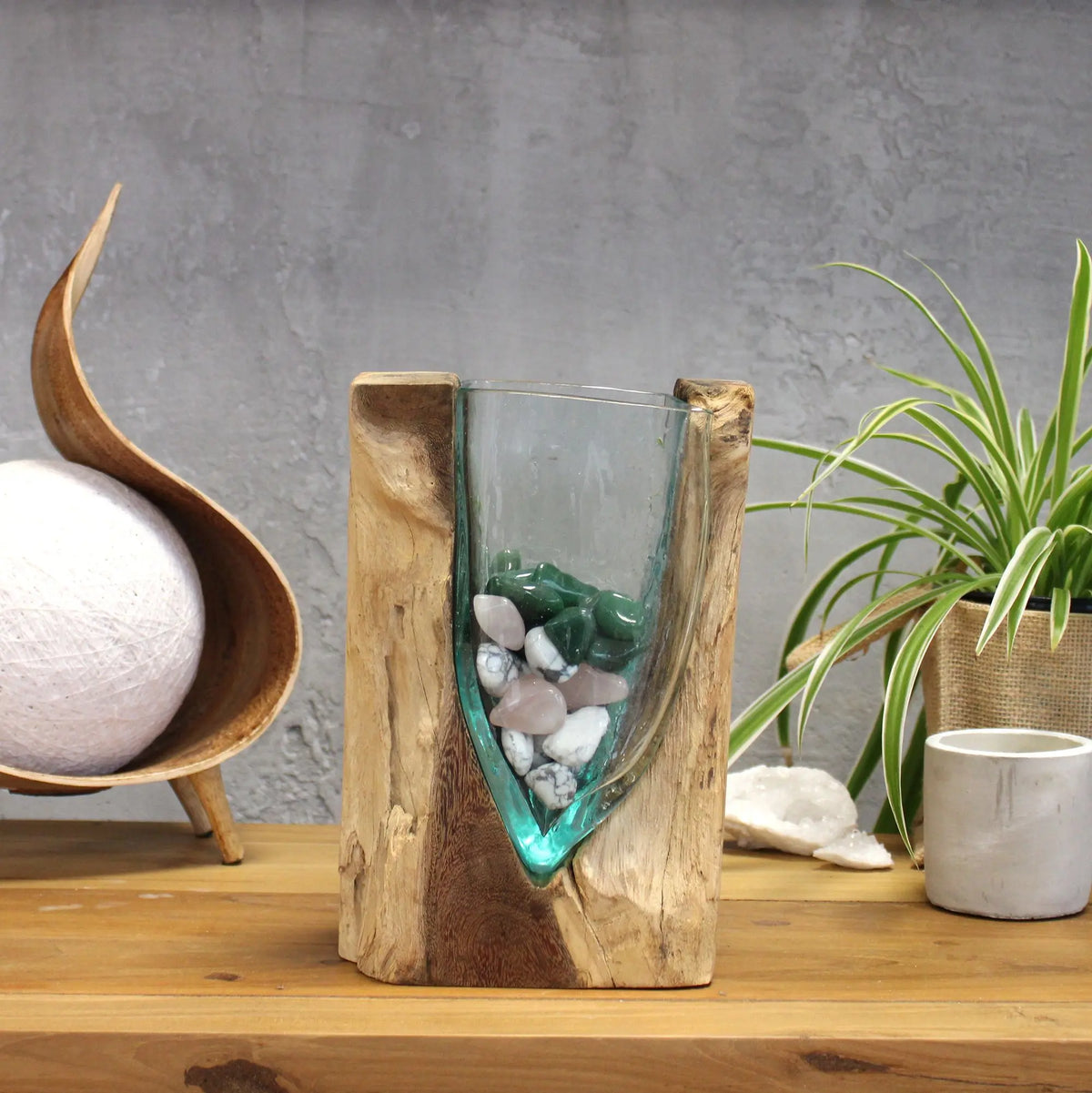 Jarrón en forma de V de vidrio fundido sobre base de madera natural, relleno con piedras decorativas de colores.