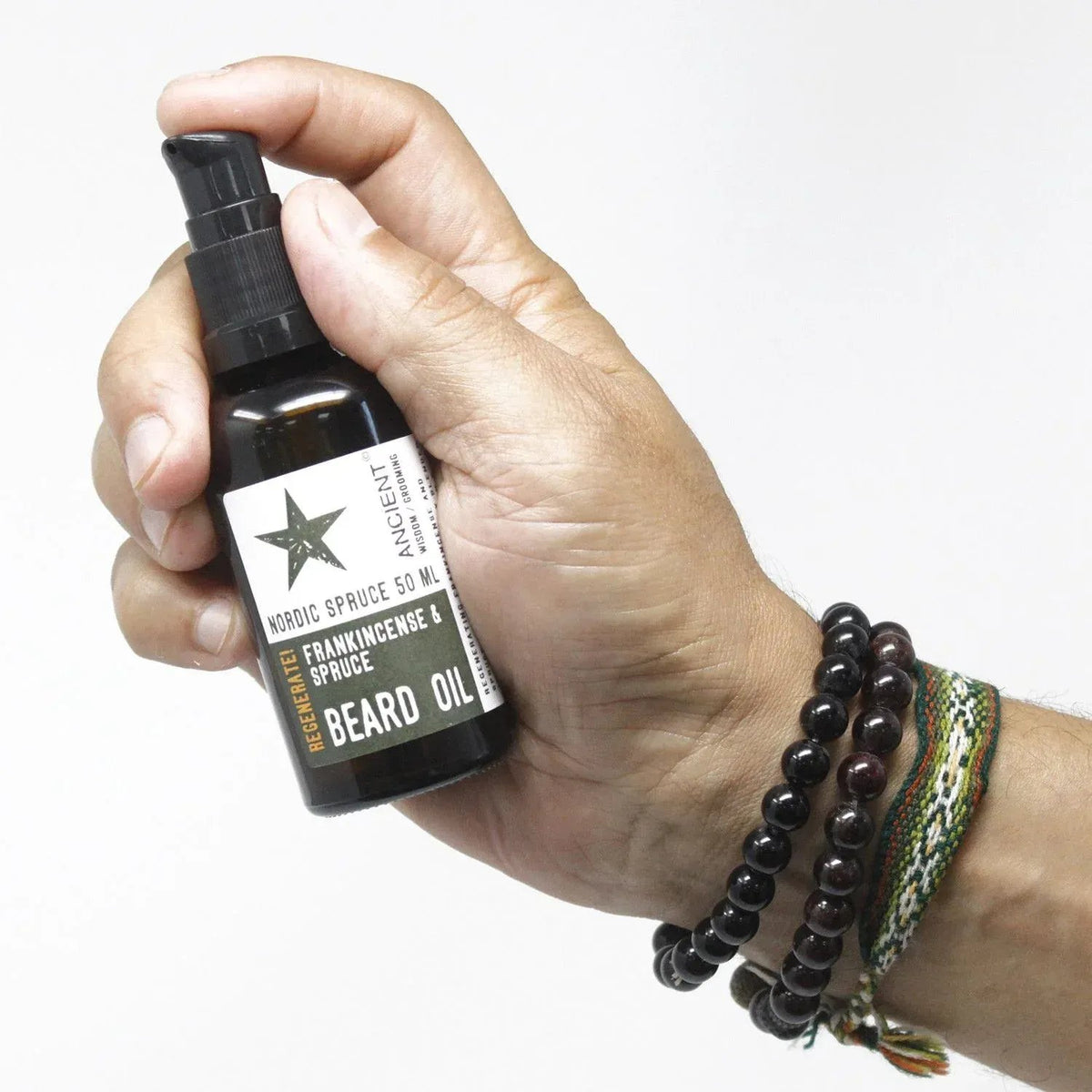 Mano sosteniendo frasco de vidrio ámbar de 50ml con dosificador de aceite para barba Ancient Wisdom sobre fondo blanco.