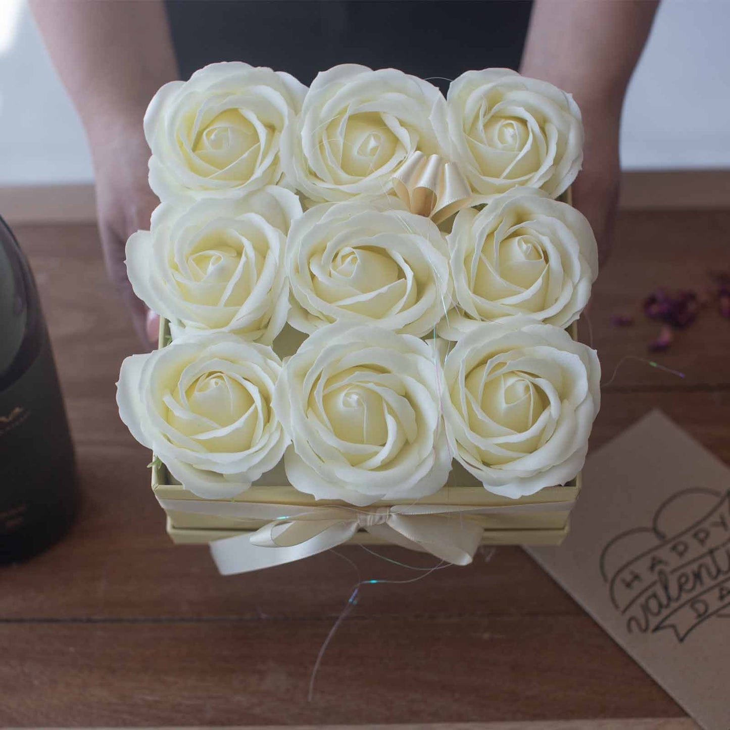 Caja cuadrada color crema con 9 rosas de jabón blancas y un lazo decorativo, sostenida por manos.
