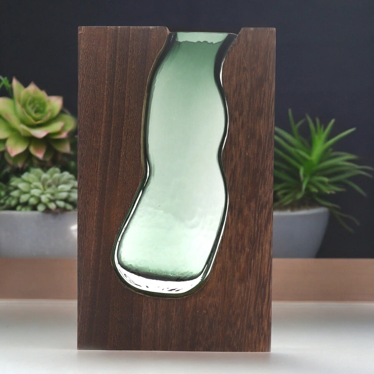 Jarrón de cristal verde con forma orgánica encajado en un bloque de madera de tung oscura de estilo natural y moderno