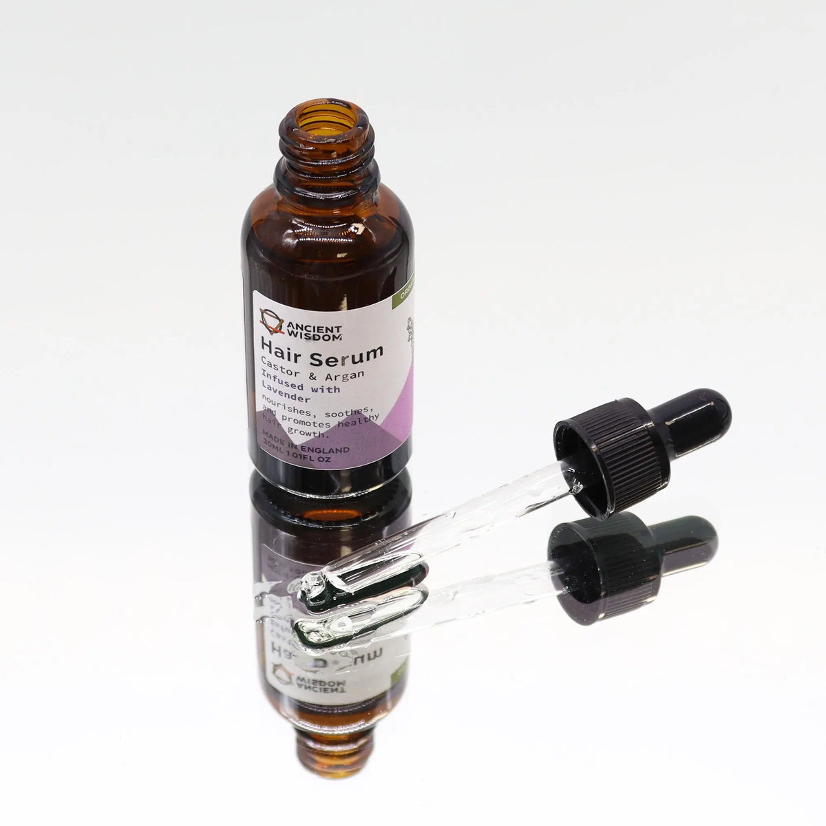 Botella de vidrio ámbar de 30ml con serum capilar y gotero sobre superficie reflectante. Etiqueta blanca y morada.