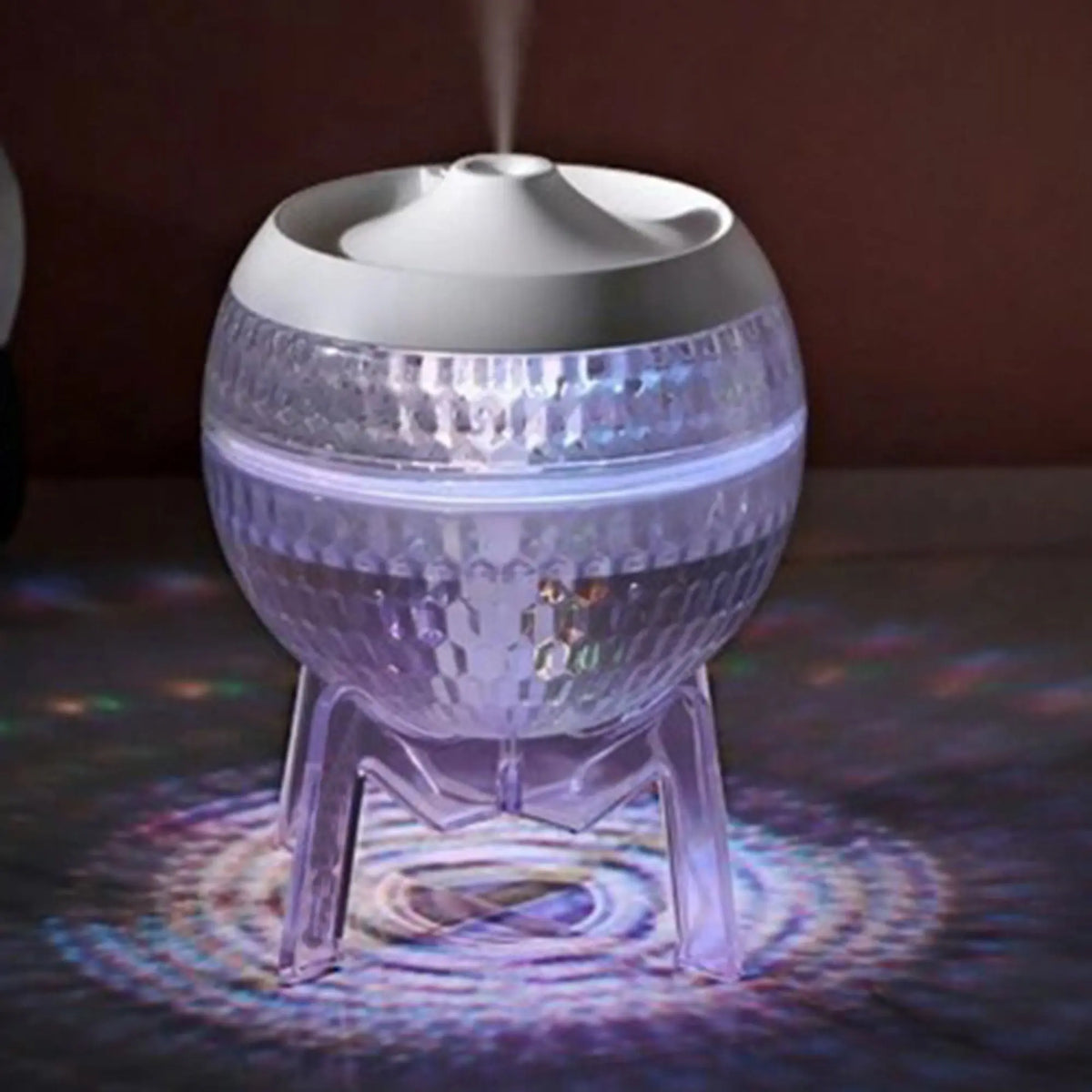 Difusor esférico transparente tipo planeta sobre soporte, emitiendo vapor y luz LED multicolor, marca Ancient Wisdom.