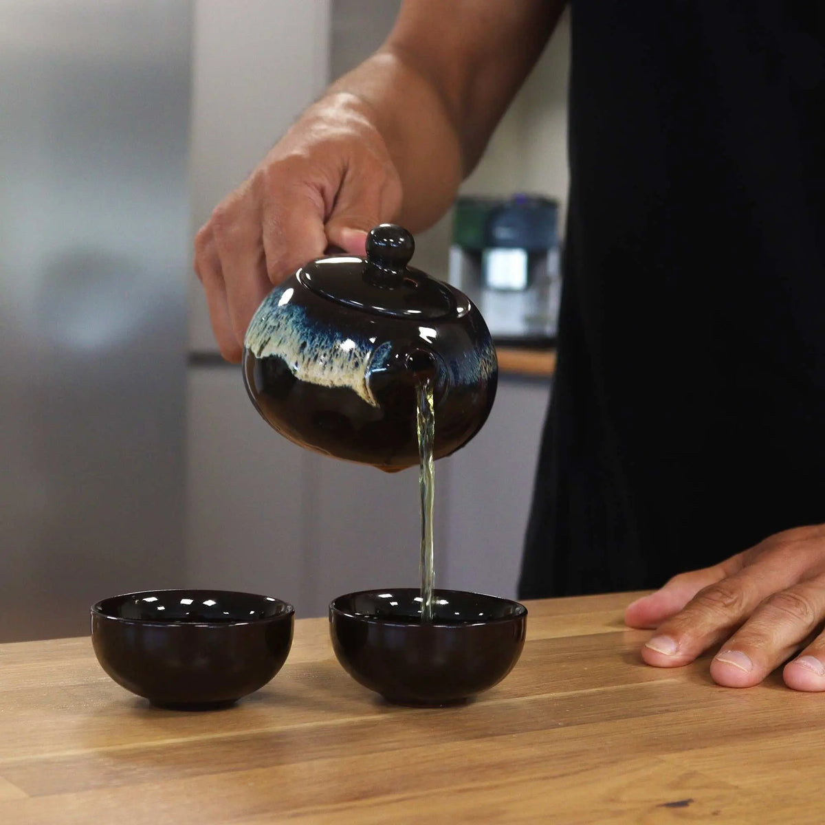 Tetera esmaltada negra con detalles azules vertiendo té en dos tazas pequeñas sobre una mesa de madera.