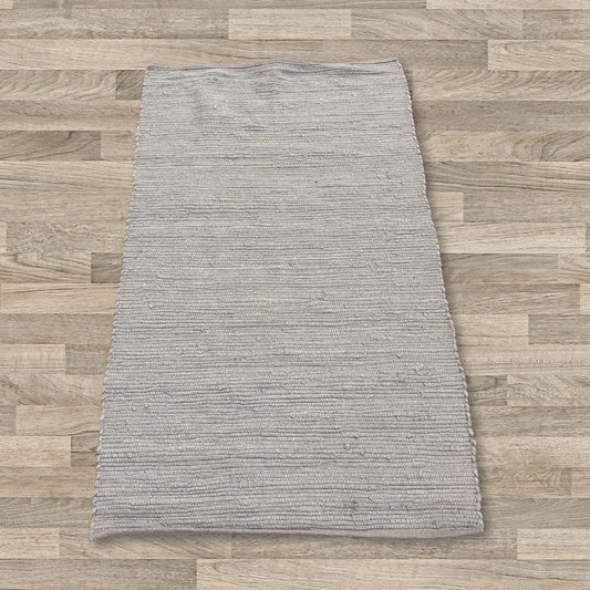 Alfombra pequeña de algodón gris, tejida a mano con textura lineal, de forma rectangular sobre un suelo de madera clara.