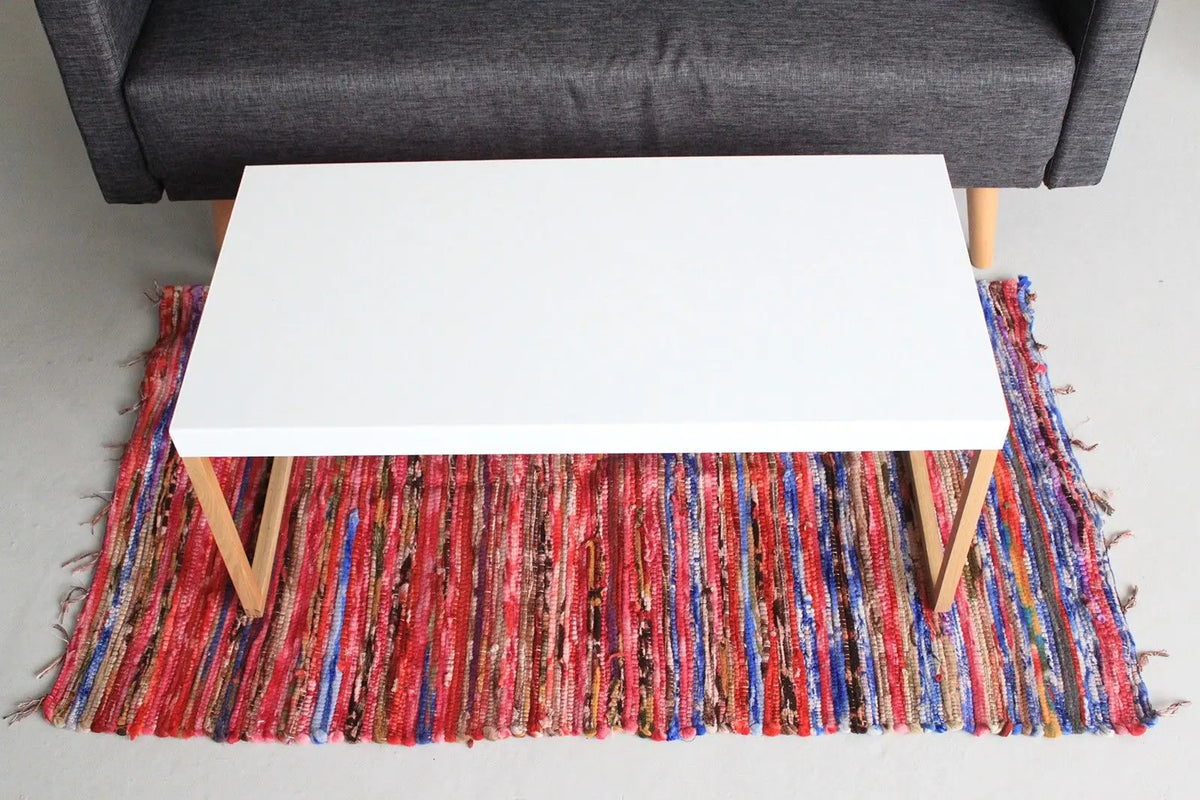 Alfombra de trapo rectangular hecha de tiras textiles multicolores, con flecos en los extremos, bajo una mesa blanca.