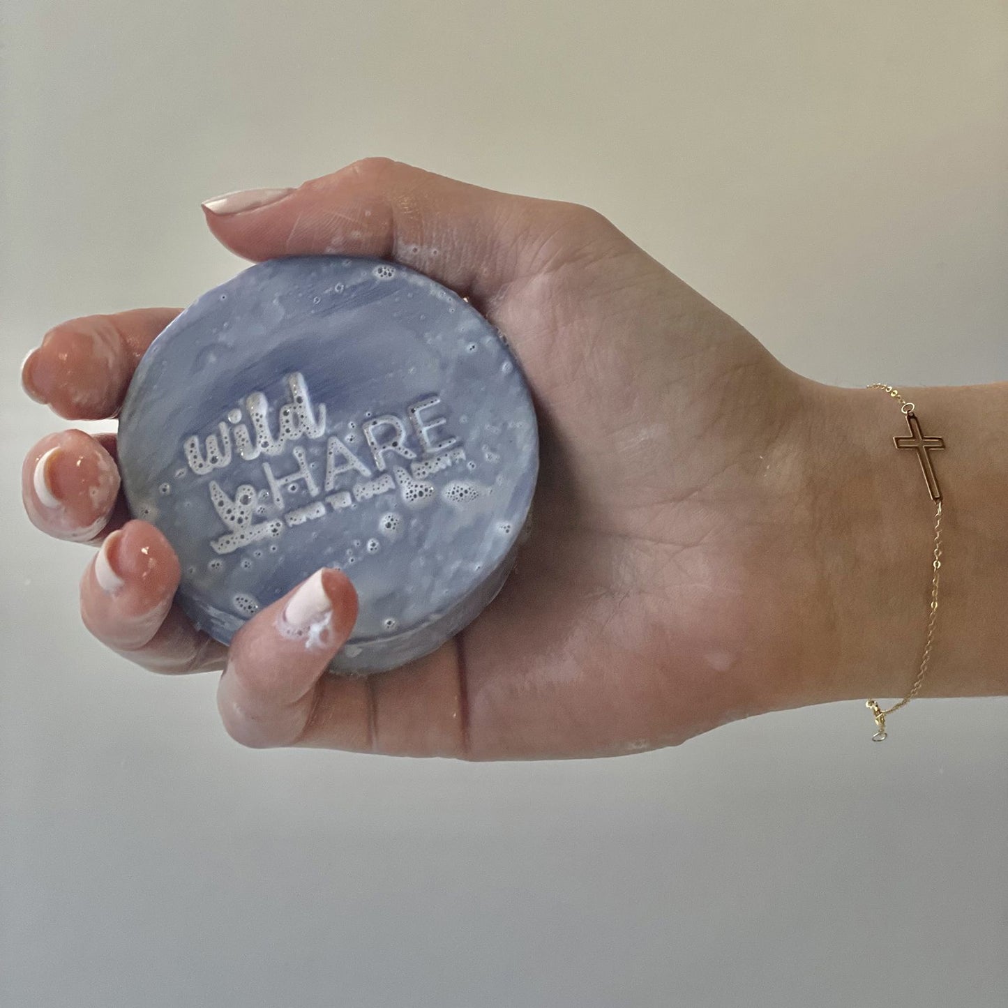 Mano sosteniendo una pastilla de champú sólida, circular y de color lila con espuma y relieve en la superficie.
