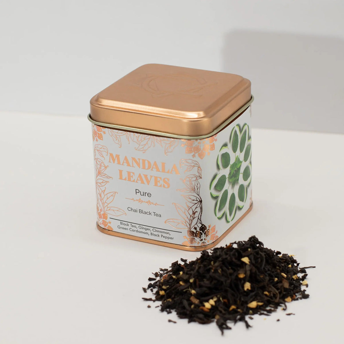 Lata cuadrada metálica color cobre con té negro chai y una pequeña pila de hojas de té sueltas sobre fondo blanco.