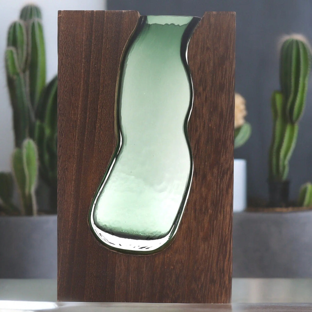 Florero de vidrio verde con forma orgánica encajado en una pieza rectangular de madera oscura de tono veteado.