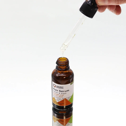 Mano sosteniendo un gotero con serum sobre una botella de vidrio ámbar de 30ml de aceite capilar. Fondo blanco.