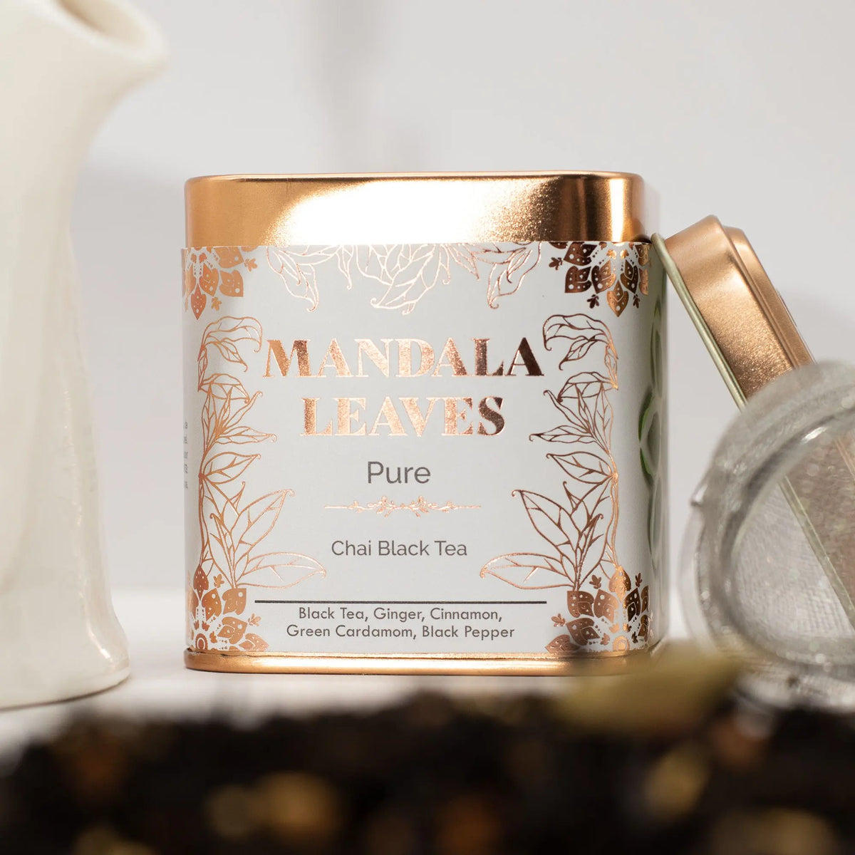 Lata metálica cuadrada de té chai, con diseño blanco y detalles en cobre, junto a un infusor sobre hojas de té.