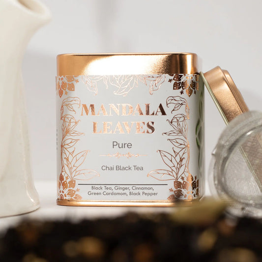 Lata metálica cuadrada de té chai, con diseño blanco y detalles en cobre, junto a un infusor sobre hojas de té.