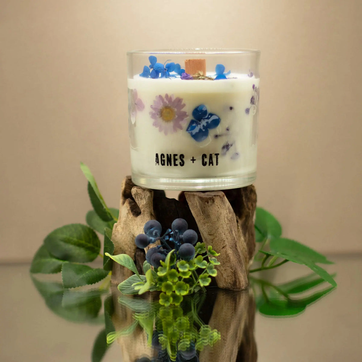 Vela aromática blanca con flores secas en vaso de vidrio, decorada sobre base de madera natural.