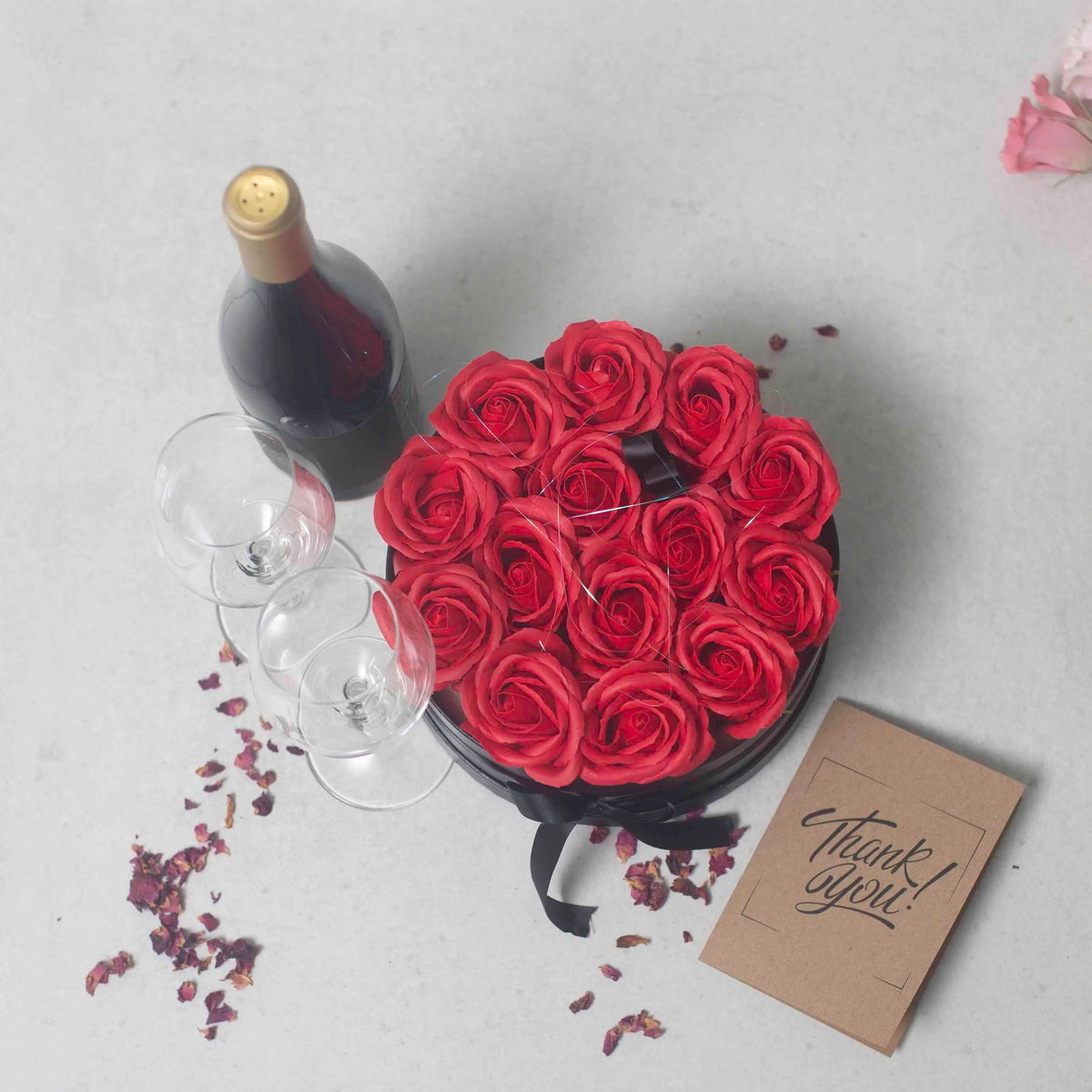 Caja redonda con 14 rosas rojas de jabón, botella de vino, dos copas y una tarjeta de agradecimiento en fondo gris.