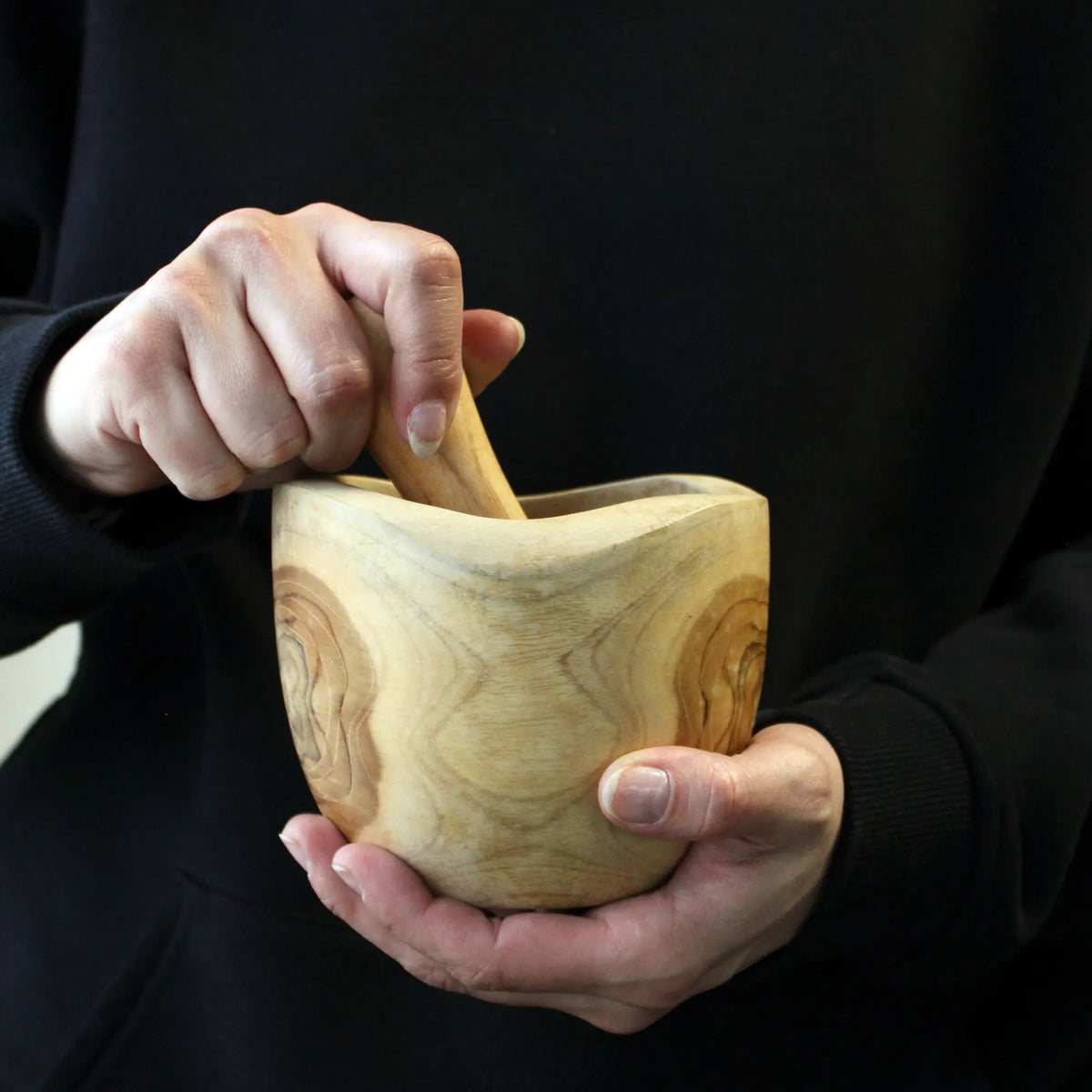 Mortero y maja de madera de teca con veta natural, sostenidos por manos contra un fondo negro. Estilo artesanal.