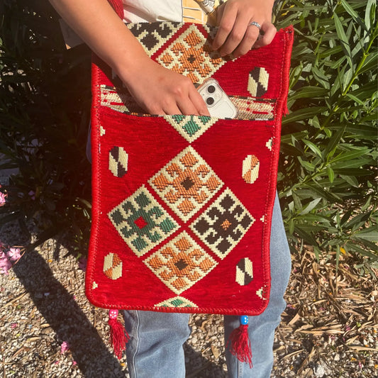 Bolso bandolera rojo de tejido kilim con patrón geométrico, detalles de borlas y bolsillo frontal con un teléfono.