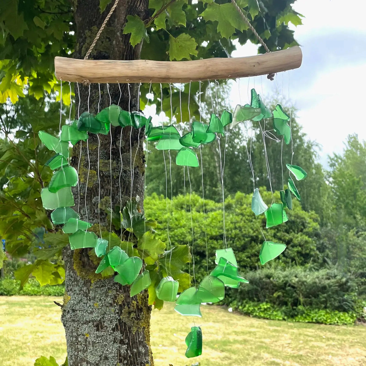 Carillón de trozos de vidrio verde colgando de una rama de madera, formando una estructura circular en un jardín.