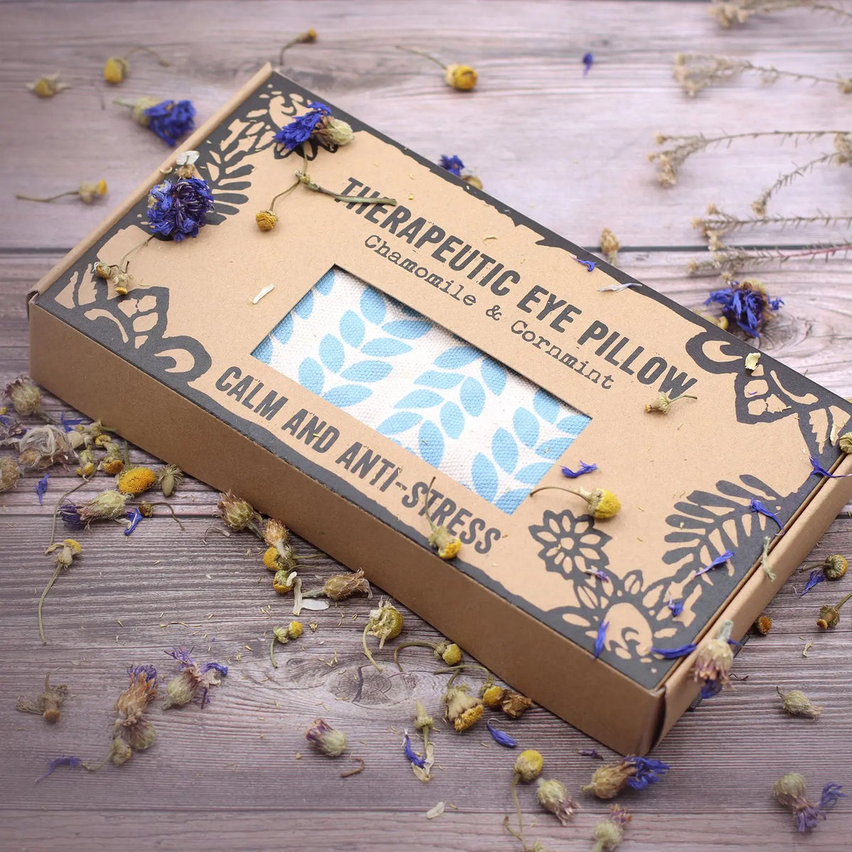 Almohada para ojos Agnes & Cat en caja de cartón decorada, con estampado de hojas azules, rodeada de flores secas.
