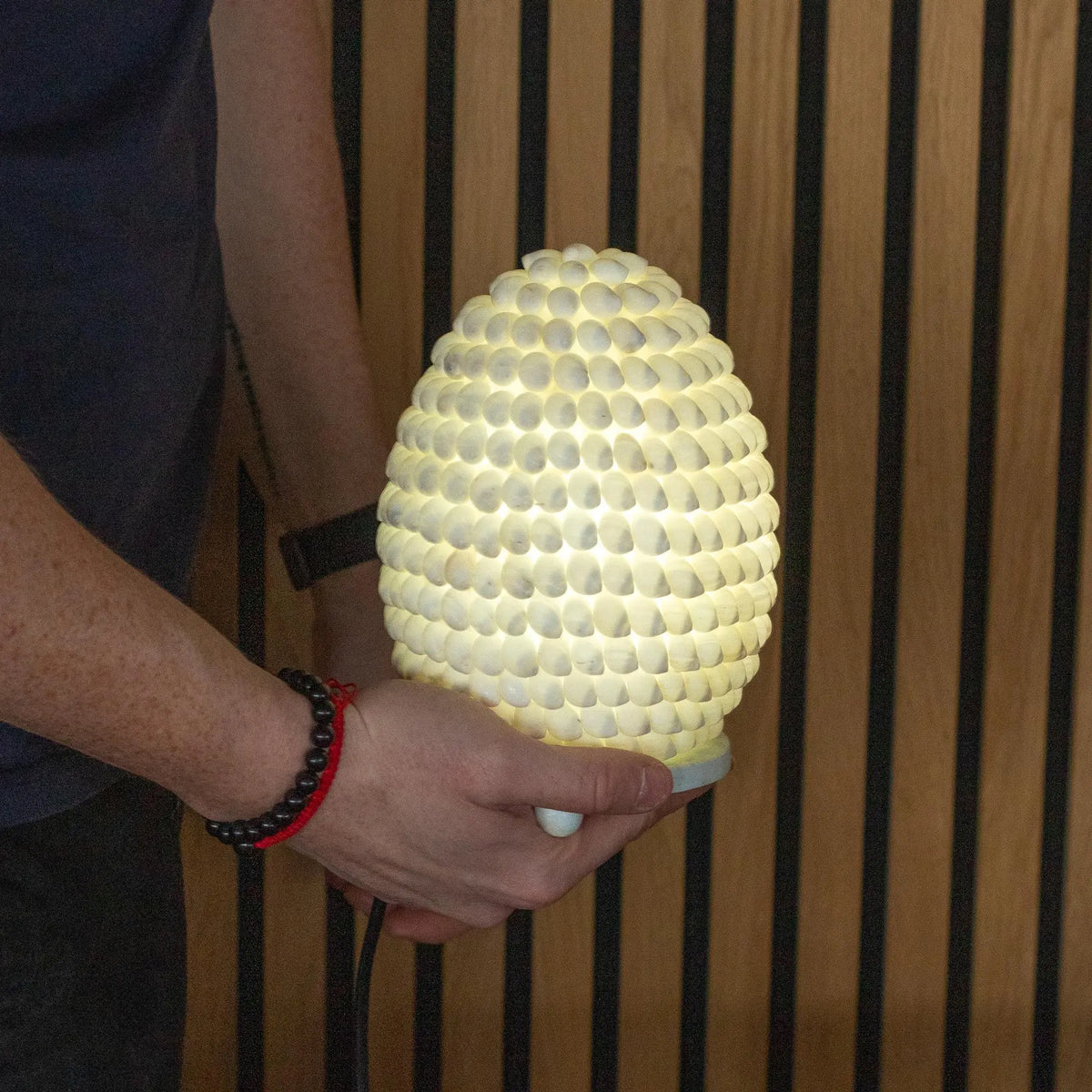 Lámpara boho ovalada de conchas marinas blancas iluminada, sostenida por unas manos, sobre fondo de listones de madera.