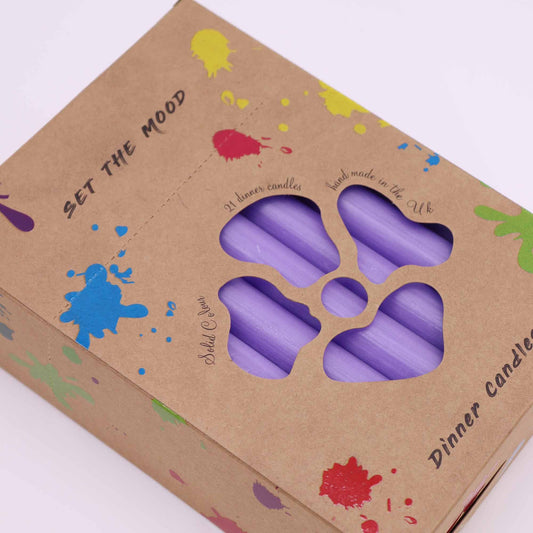 Caja de cartón con manchas de colores, mostrando a través de una abertura con forma de flor velas de mesa color lila.